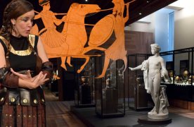 Mujer con ropas griegas, cratera con iguras griegas, y vista de la sala de Grecia con una estatua y vitrinas con cerámicas