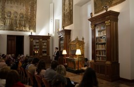 Sala con varias filas de público, un pequeño órgano dorado, y un hombre de pie cerca del órgano hablando. En la parte baja de las paredes hay muebles de biblioteca, y en la parte alta tapices