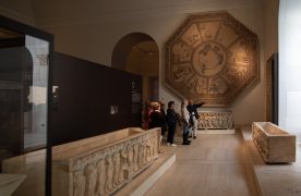 Sala de museo con tres sarcófagos de mármol sobre el suelo, un gran mosaico octogonal sobre una pared, y un grupo de visitantes