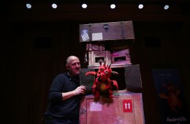 Decorado de teatral con cajas marrones con pegatinas, actor de negro, y marioneta de dragón rojo