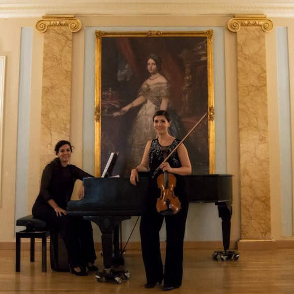 Fotografía en la que aparece una mujer joven sentada en posición de tocar un piano de colar y, delante del piano, una violinista joven con su violín. Ambas visten de negro