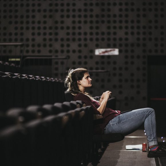 Fotografia de la Carla Rovira, l'artista resident i directora de l'espectacle. A la fotografia apareix de perfil asseguda en una butaca de teatre, mirant a l'escenari