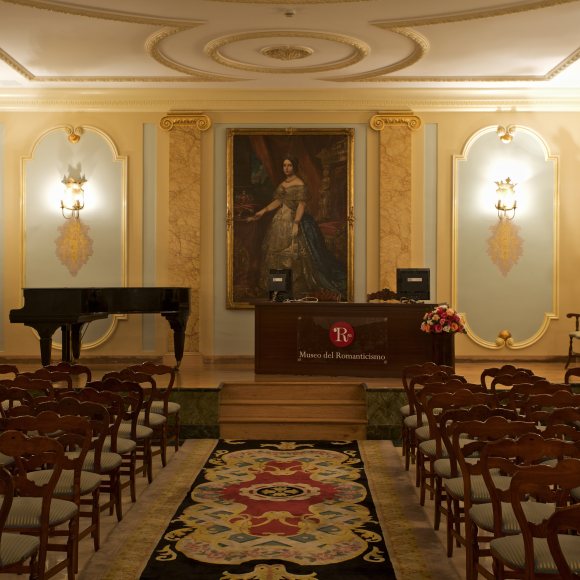 Interior del auditorio del museo. En el escenario se encuentran, a la izquierda, un piano de cola, y en el centro una mesa y una retrato de una reina. 