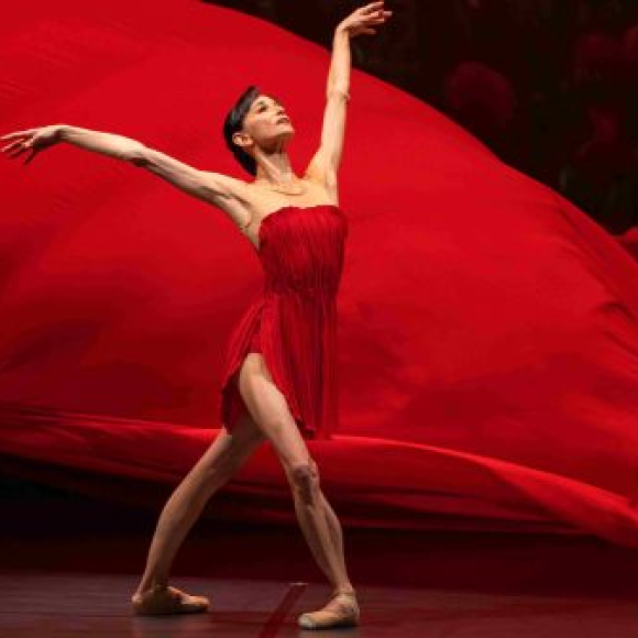 Imagen de una bailarina de ballet vestida de rojo y con una tela roja detrás en el aire.