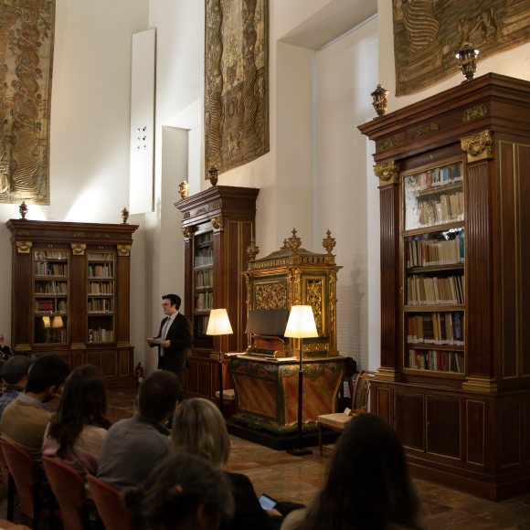 Sala con varias filas de público, un pequeño órgano dorado, y un hombre de pie cerca del órgano hablando. En la parte baja de las paredes hay muebles de biblioteca, y en la parte alta tapices