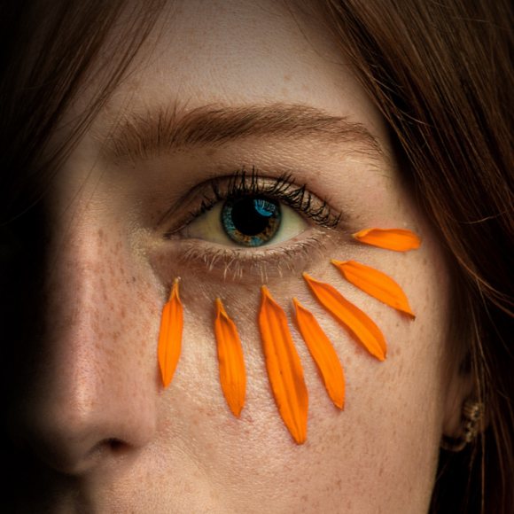 La mitad de una cara con un ojo rodeado por pétalos de flor naraja. 