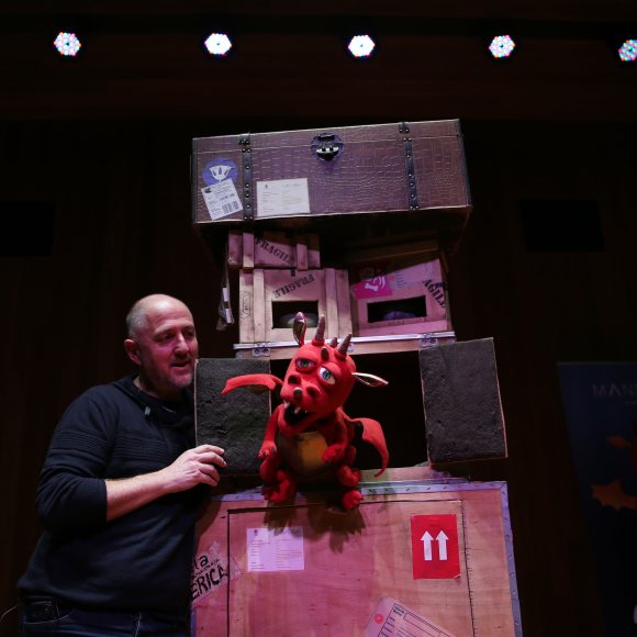 Decorado de teatral con cajas marrones con pegatinas, actor de negro, y marioneta de dragón rojo